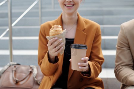 Téléchargez les photos : Plan recadré de jeune femme d'affaires souriante avec une pâtisserie appétissante fraîche et une tasse de café à emporter assis devant la caméra à la pause - en image libre de droit