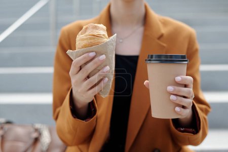 Téléchargez les photos : Mains de jeune femme d'affaires moderne tenant croissant frais et tasse de café à emporter tout en étant assis devant la caméra à la pause déjeuner - en image libre de droit