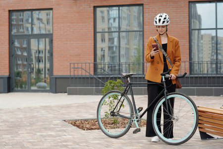 Téléchargez les photos : Jeune femme entrepreneure en casque de sécurité et vêtements décontractés défilant dans le téléphone portable tout en se tenant à côté de son vélo contre le bâtiment moderne - en image libre de droit