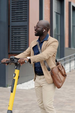 Photo for Young successful businessman in quiet luxury attire and earphones holding by handles of scooter while listening to music in urban environment - Royalty Free Image