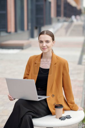 Photo for Young successful female student or freelancer in quiet luxury attire looking at camera while sitting in urban environment and using laptop - Royalty Free Image