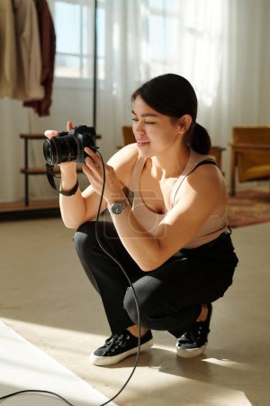 Photo for Young smiling female photographer with photocamera sitting on squats in front of camera while taking pictures of model during photo session - Royalty Free Image