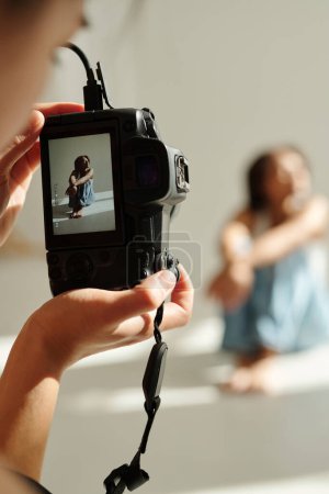 Téléchargez les photos : Focus sur l'écran de la photocaméra tenue par une jeune photographe professionnelle prenant des photos de mannequin assis en studio - en image libre de droit