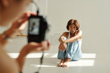 Téléchargez les photos : Jeune jolie mannequin de mode afro-américaine en casual posant pour appareil photo tout en étant assis devant le photographe pendant la séance photo - en image libre de droit
