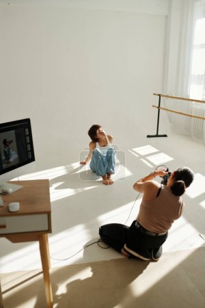 Téléchargez les photos : Jeune femme afro-américaine en jeans bleus assise sur le sol par un mur blanc tandis que le photographe professionnel prend des photos d'elle en studio - en image libre de droit