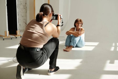 Téléchargez les photos : Bonne fille afro-américaine en tenue décontractée posant pour appareil photo tout en étant assis sur le sol par le mur devant le photographe lors de la séance photo - en image libre de droit