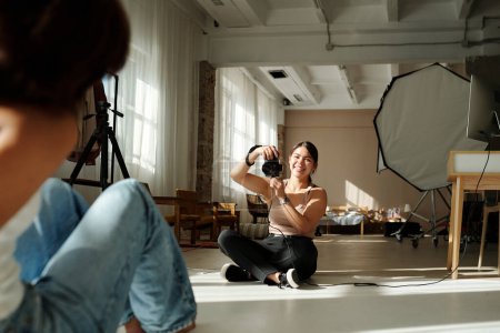 Téléchargez les photos : Photographe souriante avec appareil photo assis dans un studio spacieux en face du mannequin de mode en tenue décontractée et prendre des photos d'elle - en image libre de droit