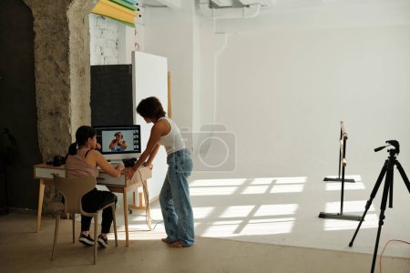 Téléchargez les photos : Deux jeunes femmes interculturelles en tenue décontractée discutent de nouveaux plans à l'écran d'ordinateur après une séance photo alors que l'une d'elles est assise sur une chaise - en image libre de droit