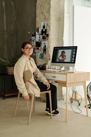 Téléchargez les photos : Jeune photographe professionnel regardant la caméra tout en étant assis sur le lieu de travail avec écran d'ordinateur avec des photos de modèle de mode à l'écran - en image libre de droit