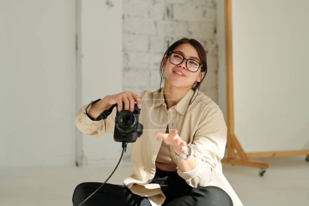 Téléchargez les photos : Joyeux jeune photographe en lunettes et vêtements décontractés tenant un appareil photo assis sur le sol du studio et parlant au mannequin - en image libre de droit