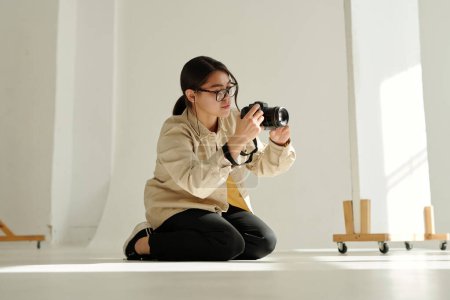 Téléchargez les photos : Jeune photographe avec appareil photo debout à genoux sur le sol du studio et prenant des photos de mannequin pendant la séance photo - en image libre de droit