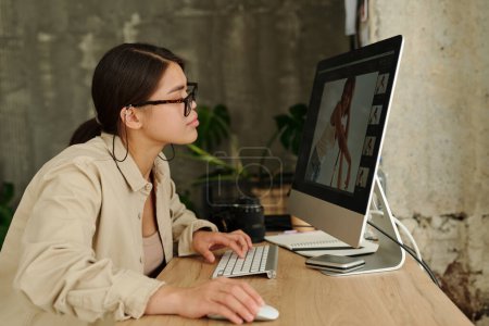 Téléchargez les photos : Vue latérale de la jeune femme en tenue décontractée regardant à travers de nouveaux clichés de mannequin à l'écran de l'ordinateur après la séance photo en studio - en image libre de droit