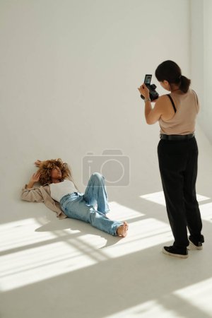 Téléchargez les photos : Jeune femme afro-américaine en tenue décontractée allongée par terre au mur pendant la séance photo tout en posant pour une photographe en studio - en image libre de droit