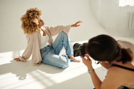 Téléchargez les photos : Jeune jolie mannequin féminine en tenue décontractée assise sur le sol du studio contre le mur et posant devant un photographe professionnel - en image libre de droit