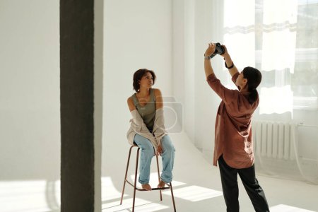 Téléchargez les photos : Jeune photographe prenant au-dessus de l'angle photo du mannequin de mode en tenue décontractée assis devant la caméra et posant pendant la séance photo - en image libre de droit