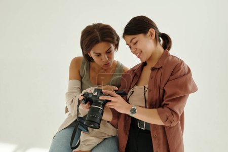 Téléchargez les photos : Joyeux jeune photographe avec appareil photo montrant de nouvelles photos de mannequin tout en se tenant en studio contre un mur blanc - en image libre de droit