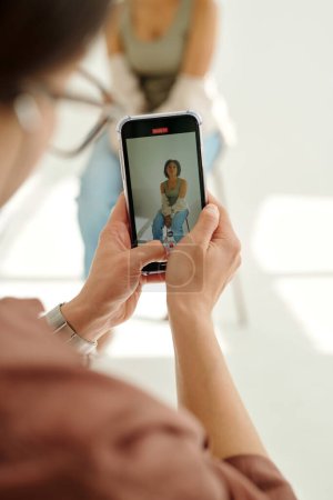 Téléchargez les photos : Focus sur l'écran du smartphone avec prise de vue de jeune mannequin de mode féminine dans les mains du photographe prenant des photos sur gadget mobile en studio - en image libre de droit