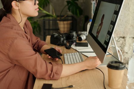 Téléchargez les photos : Plan recadré d'une jeune photographe avec smartphone assis devant l'écran de l'ordinateur et examinant de nouvelles images de mannequin - en image libre de droit