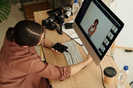 Téléchargez les photos : Au-dessus de l'angle du jeune photographe indépendant assis devant l'écran de l'ordinateur avec de nouvelles photos de mannequin prises lors de la séance photo - en image libre de droit
