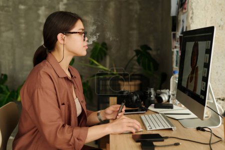 Photo for Side view of young brunette woman smoking e-cigarette while sitting in front of computer screen with new shots of fashion model during review - Royalty Free Image