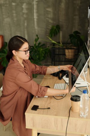Téléchargez les photos : Vue latérale du jeune photographe réglant le moniteur d'ordinateur tout en étant assis par le lieu de travail en studio et faisant l'examen de nouveaux clichés - en image libre de droit
