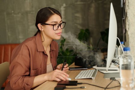 Téléchargez les photos : Jeune femme avec cigarette électronique regardant l'écran d'ordinateur tout en examinant de nouveaux plans de modèle et en choisissant les plus dignes - en image libre de droit