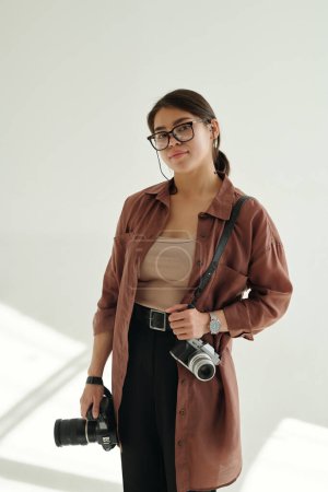 Photo for Young stylish photographer with two photocameras looking at you through eyeglasses while standing in photo studio in isolation - Royalty Free Image