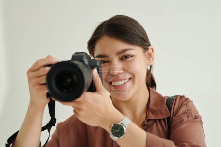 Téléchargez les photos : Joyeux jeune photographe avec appareil photo prenant des photos isolées pendant la séance photo en studio et vous regardant avec sourire - en image libre de droit