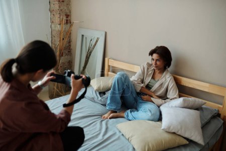 Téléchargez les photos : Focus sur jeune mannequin femme assise sur un lit double et regardant la caméra tenue par le photographe prendre des photos pendant la séance photo - en image libre de droit