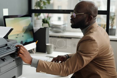 Téléchargez les photos : Jeune homme d'affaires dans les lunettes et les vêtements de coffrage appuyant sur le bouton de démarrage sur le panneau de l'imprimante tout en étant assis par le lieu de travail devant l'écran d'ordinateur - en image libre de droit