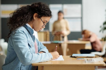 Téléchargez les photos : Vue latérale d'une écolière sérieuse portant des lunettes prenant des notes dans un copybook alors qu'elle était assise à son bureau à la leçon de grammaire de la langue anglaise - en image libre de droit