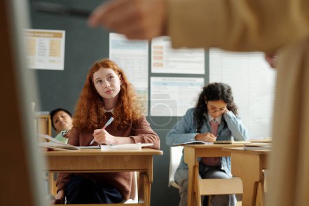 Téléchargez les photos : Écolière intelligente avec stylo écoutant l'explication de l'enseignant et prenant des notes dans le copybook tout en étant assise par bureau parmi ses camarades de classe - en image libre de droit