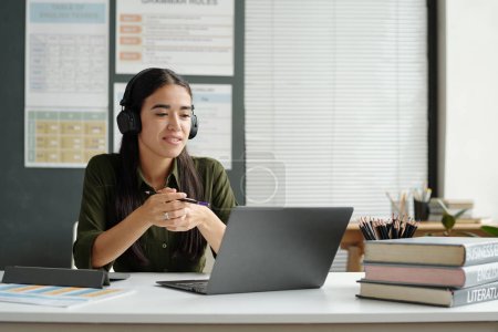 Téléchargez les photos : Jeune professionnel confiant parlant à un apprenant en ligne sur un écran d'ordinateur portable assis à son bureau en classe avec des tables de grammaire au mur - en image libre de droit