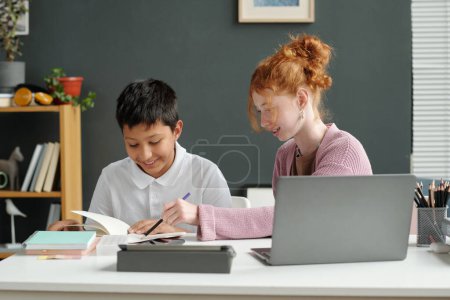 Téléchargez les photos : Écolière intelligente pointant vers la page de copybook ouvert de son camarade de classe tout en étant assis à côté de lui pendant la leçon en ligne devant l'ordinateur portable - en image libre de droit