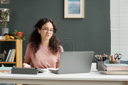 Photo for Cute secondary school learner in earphones talking to teacher or answering questions while looking at laptop screen during online lesson - Royalty Free Image