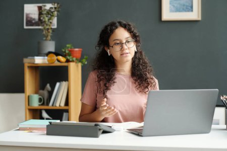 Téléchargez les photos : Écolière intelligente en t-shirt rose regardant l'écran de l'ordinateur portable et répondant à la question de l'enseignant pendant la leçon en ligne de langue anglaise - en image libre de droit