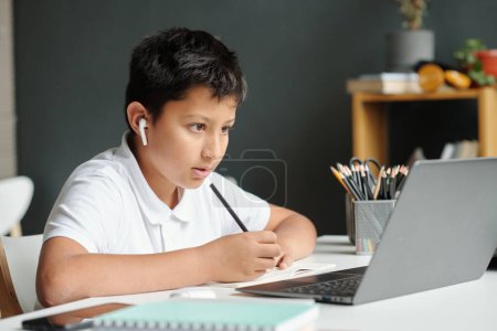 Foto de Colegial multiétnico serio con lápiz mirando la pantalla del ordenador portátil mientras escucha al profesor durante la lección en línea y toma notas en un libro de copas - Imagen libre de derechos