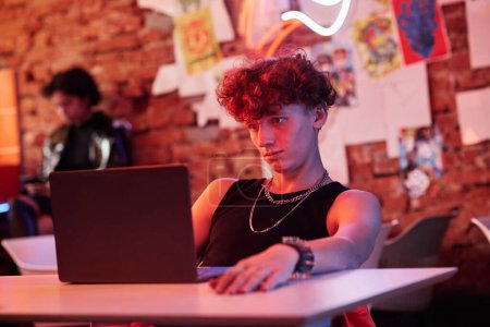 Téléchargez les photos : Un gars sérieux en tenue décontractée regardant un écran d'ordinateur portable assis dans un bar ou un café cyberpunk et jouant à un jeu virtuel ou regardant une vidéo en ligne - en image libre de droit