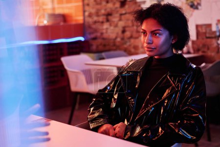 Photo for Young African American woman wearing black shiny varnished leather jacket sitting in armchair by table in front of her boyfriend in cafe - Royalty Free Image