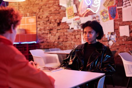 Photo for African American girl in black shiny varnished leather jacket sitting by table in front of her boyfriend in cyberpunk cafe and looking at him - Royalty Free Image