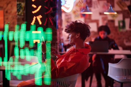 Foto de Vista lateral del joven en chaqueta de bombardero desplazándose en la pantalla de la tableta mientras está sentado en el bar cyberpunk o cafetería con luces de neón y colores vibrantes - Imagen libre de derechos