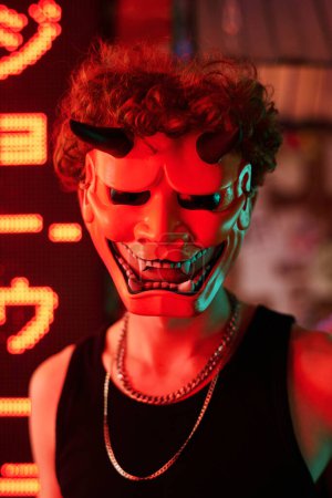 Foto de Personaje malvado en máscara de pie contra el jeroglífico que significa mejor ramen y mirando a la cámara en el club cyberpunk iluminado por luces de neón - Imagen libre de derechos