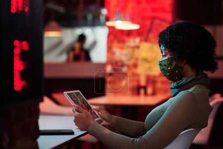 Foto de Vista lateral de mujer joven en mascarilla facial escalofriante en cyberpunk café y mirando la pantalla de la tableta mientras ve video en línea o jugando - Imagen libre de derechos