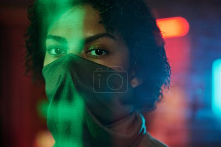 Foto de Cara de chica afroamericana medio cubierta con máscara mirando a la cámara mientras está de pie en el bar cyberpunk con luces de neón en el fondo - Imagen libre de derechos