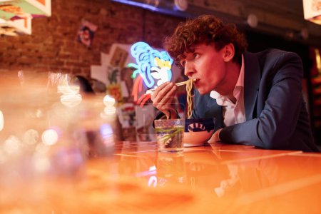 Photo for Young hungry businessman in formalwear eating Japanese ramen for lunch in cyberpunk bar, cafe or restaurant with neon lights or illuminations - Royalty Free Image