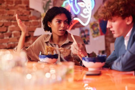 Foto de Joven mujer afroamericana hablando con su colega o novio mientras está sentada frente a él y ambos comiendo ramen en el almuerzo en la cafetería - Imagen libre de derechos