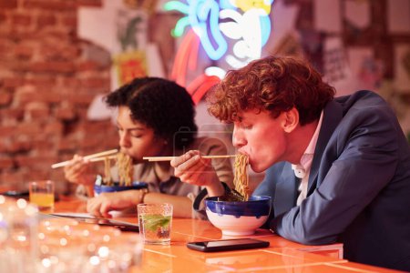 Foto de Joven hombre hambriento con el pelo rizado inclinado sobre la mesa con tazón mientras come sabroso ramen cocinado por el chef de la cafetería japonesa o restaurante - Imagen libre de derechos