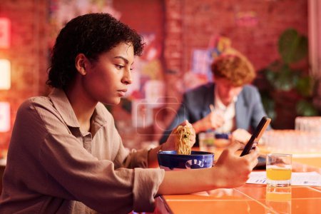 Foto de Chica seria con teléfono celular mirando la pantalla del gadget mientras está sentado en la cafetería japonesa, teniendo ramen y viendo vídeo en línea o mensajes de texto - Imagen libre de derechos