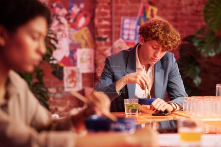 Téléchargez les photos : Focus sur les jeunes hommes d'affaires en tenue de cérémonie mangeant des ramen avec des baguettes assis à table dans un café japonais ou un bar avec un design cyberpunk - en image libre de droit