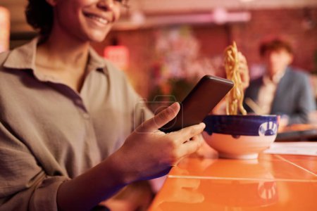 Foto de Enfoque en el teléfono inteligente en la mano de la mujer joven que tiene comida japonesa en la cafetería o el restaurante y la comunicación en el chat de vídeo o mensajes de texto - Imagen libre de derechos